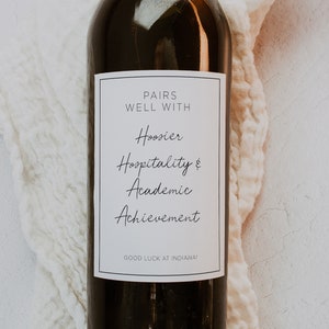 May include: A bottle of wine with a white label that says "Pairs Well With Hoosier Hospitality & Academic Achievement. Good Luck at Indiana!"