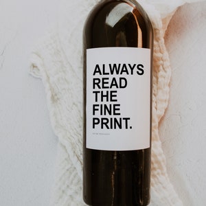 May include: A dark wine bottle with a white label featuring the text "ALWAYS READ THE FINE PRINT." The bottle is set against a white, textured cloth. The words "WE'RE PREGNANTI" are at the bottom of the label.
