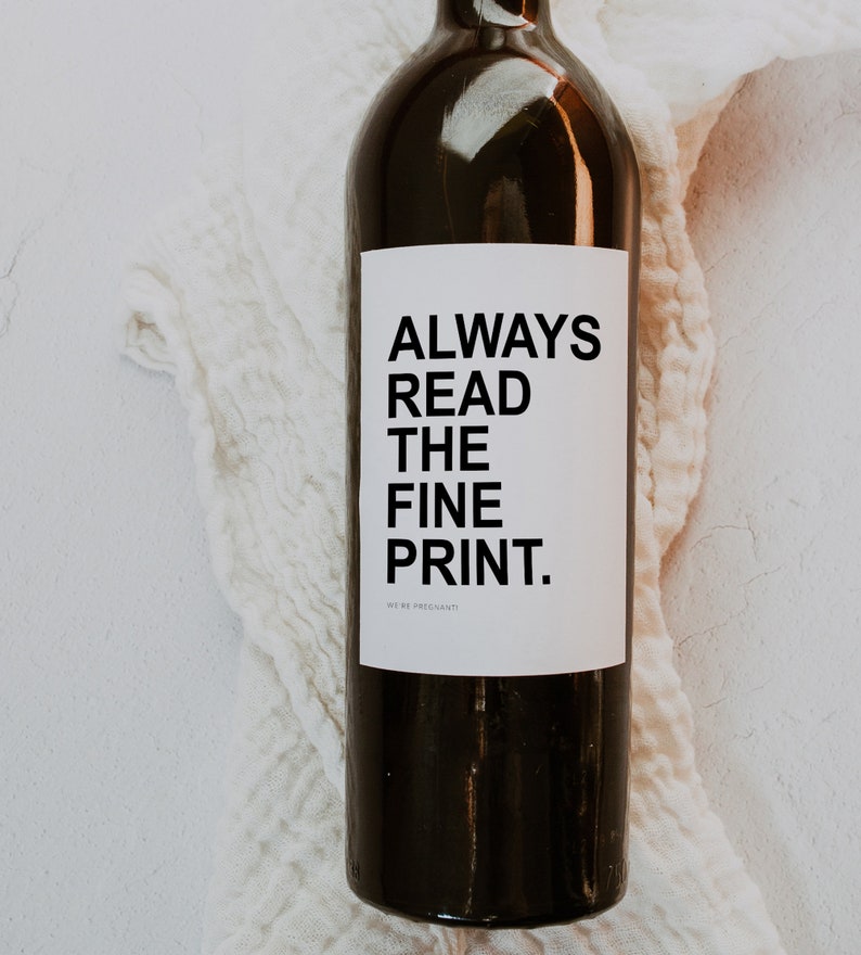 May include: A dark wine bottle with a white label featuring the text "ALWAYS READ THE FINE PRINT." The bottle is set against a white, textured cloth. The words "WE'RE PREGNANTI" are at the bottom of the label.
