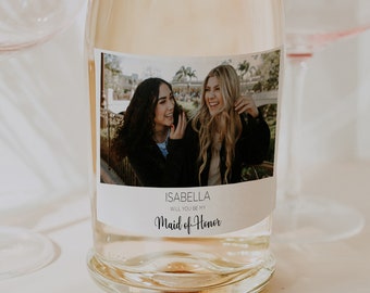 Peut inclure: Une bouteille de champagne transparente avec une étiquette portant une photo de deux femmes souriantes. L'étiquette indique "ISABELLA WILL YOU BE MY Maid of Honor". Deux verres à champagne avec des bords dorés sont en arrière-plan.