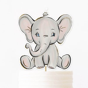 Puede incluir: Un adorno para pastel de elefante gris con ojos azules y detalles de purpurina dorada. El elefante está sentado con la trompa enroscada y las orejas levantadas.