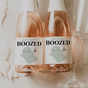 Può includere: Due bottiglie di vino spumante rosato con etichette bianche che recitano "you've been BOOZE'D" e raffigurano un fantasma dei cartoni animati che tiene una bottiglia di liquido viola.