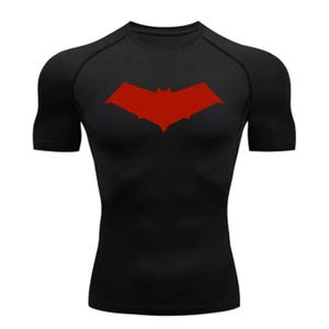 Puede incluir: Camiseta de compresión negra de manga corta con un logotipo de murciélago rojo en la parte delantera.