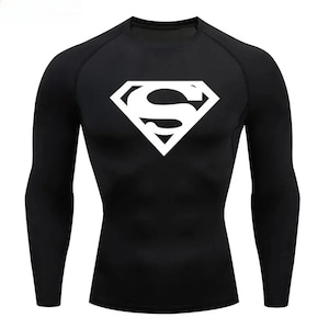 Op de afbeelding: Zwart compressieshirt met lange mouwen en een wit Superman-logo op de borst. Het logo is een witte "S" in een ruit. Het shirt heeft een ronde hals en is ontworpen voor sport.