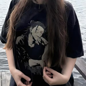 May include: Black t-shirt featuring a black and white anime-style illustration of a character with long hair and an earring. The shirt also has Japanese text. The t-shirt has short sleeves.