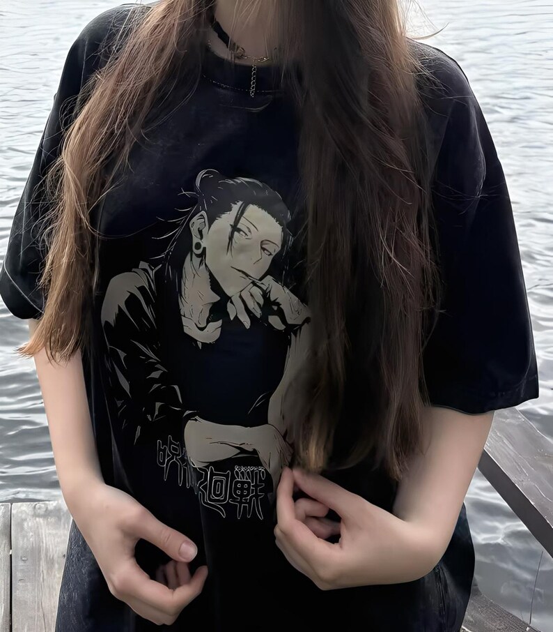 May include: Black t-shirt featuring a black and white anime-style illustration of a character with long hair and an earring. The shirt also has Japanese text. The t-shirt has short sleeves.