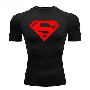 Op de afbeelding: Zwart compressieshirt met korte mouwen en een rood Superman-logo op de borst. Het logo is een rode ruit met een rode "S" erin. Het shirt heeft een ronde hals en is nauwsluitend.