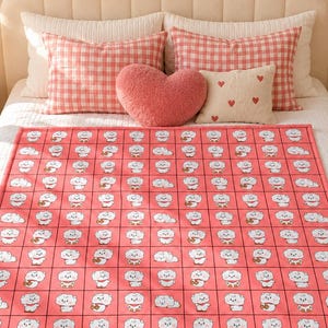 May include: A coral pink throw blanket featuring a repeating grid pattern of small white cartoon characters. Each square contains a different pose of the same white character with a red scarf, arranged in a uniform layout across the fabric.