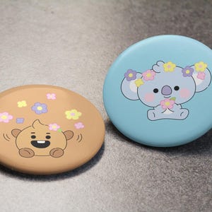 Op de afbeelding: Twee ronde buttons. De linker button is beige met een bruin stripfiguurtje omringd door kleine roze, gele en paarse bloemetjes. De rechter button is lichtblauw met een grijs koala-figuurtje dat een bloemenkrans draagt.