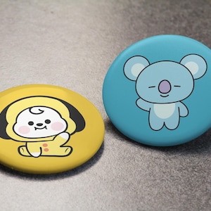 Op de afbeelding: Twee ronde, kleurrijke buttonspins. Eén is geel met een stripfiguur in een gele outfit. De andere is lichtblauw met een cartoonkoala. Beide hebben zwarte contouren en liggen op een grijze ondergrond.
