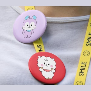 Op de afbeelding: Twee ronde buttons bevestigd aan een geel koord met het woord SMILE en smiley-icoontjes die herhaaldelijk zijn afgedrukt. De ene button is lichtpaars met een stripfiguur, de andere is rood met een witte stripfiguur.