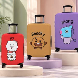 May include: Three upright suitcases with protective covers featuring BT21 characters. The red cover displays RJ, the tan cover features Shooky, and the purple cover shows Mang. Each suitcase has a retractable handle and four black wheels.