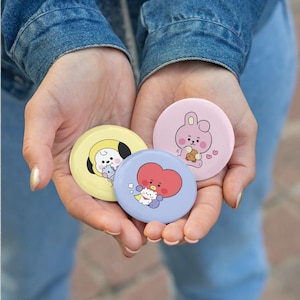 Op de afbeelding: Drie ronde buttons met stripfiguren. Eén button is geel met een wit en zwart figuurtje, één is lichtroze met een roze konijnfiguurtje en één is lichtblauw met een rood hartvormig figuurtje.