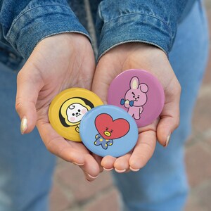 Op de afbeelding: Drie ronde buttons met cartoonfiguren. Een gele button toont een wit figuurtje met een capuchon, een blauwe button toont een rood hartvormig figuurtje en een roze button toont een roze konijn dat een blauwe halter vasthoudt.