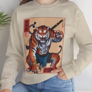 May include: A beige sweatshirt with a graphic print of a tiger holding a sword. The tiger is in a fighting stance and has a fierce expression. The print is surrounded by a red and gold border. The text "Don't underestimate the power of a tiger" is printed below the image.