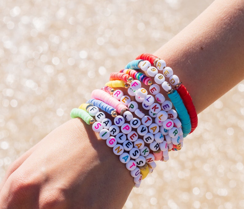 May include: A stack of colorful beaded bracelets with white letter beads spelling out words like "Fearless", "Speak", "Signature", and "1988".