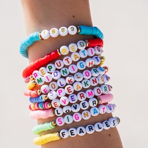 May include: A stack of colorful beaded bracelets with words spelled out on each one. The words include "Fearless", "Signature", "Lover", "Evermore", "Speak", "Folklore", "Midnight", "Reputation", "Red", and "1983".