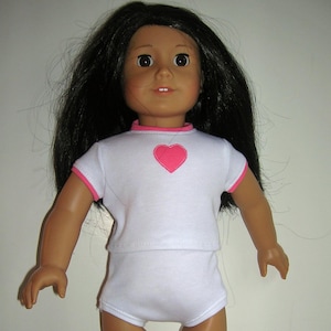 May include: A doll with long black hair and brown eyes wearing a white outfit. The outfit includes a short-sleeved top with a pink heart and trim, and matching shorts. The doll has a fair complexion.