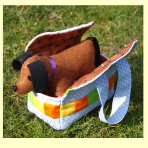 Peut inclure: Un jouet en peluche représentant un chien teckel marron avec des oreilles noires et un collier violet est placé dans un sac coloré. Le sac a un extérieur à pois bleus, un intérieur marron avec des empreintes de pattes et une poignée. Le sac est sur de l'herbe verte.