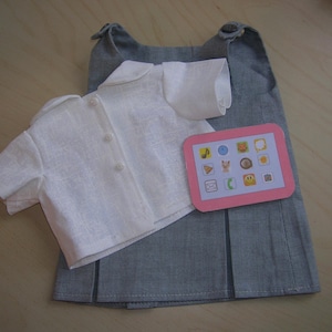 Peut inclure: Une blouse blanche à boutons et une jupe grise avec une tablette rose comportant différentes icônes. La jupe est plissée.