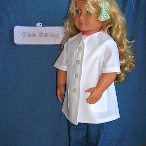 May include: A blonde doll wearing a white lab coat with buttons and a blue jean skirt. The doll is standing in front of a blue background with a sign that says "The Salon".