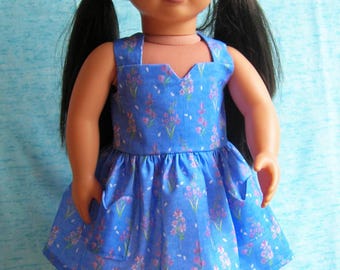 May include: A doll with long black hair styled in two pigtails, wearing a sleeveless blue dress featuring a small pink and purple floral pattern, a notched neckline, and two front pockets on the gathered skirt, standing on a white base.