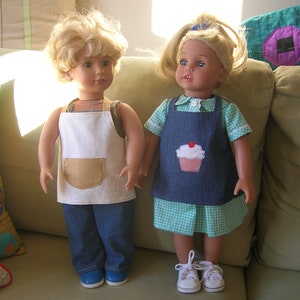 May include: Two dolls wearing aprons, one with a white apron and the other with a blue denim apron with a cupcake design. The dolls are standing on a couch with pillows behind them.