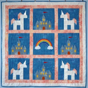 May include: A handmade quilt with a whimsical design. The quilt features a blue background with nine squares, each containing a unicorn, castle, or rainbow. The unicorns are white, and the castles are gray with yellow windows. The rainbow is multi-colored.