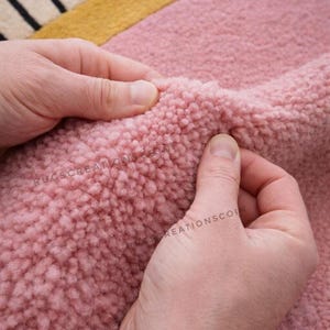 Może przedstawiać: Zbliżenie na r&oacute;żowy, teksturowany dywan. Dywan ma miękki, pluszowy wygląd. Na dywanie widoczne są słowa "RUGS CREATION" i "REATIONSCO". Dywan ma ż&oacute;łto-czarną obw&oacute;dkę w paski.