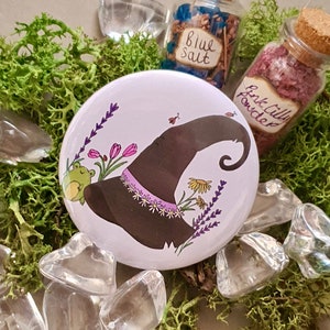 May include: A white button featuring a cartoon illustration of a witch's hat with a frog perched on the brim. The hat is adorned with flowers and lavender. The button is surrounded by clear crystals and green moss.