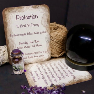 May include: A small glass jar with a purple lid filled with herbs and crystals. The jar is on a card with the text "Protection To Bind An Enemy" and instructions for use. The card is on a brown surface with a ball of twine and a black orb.