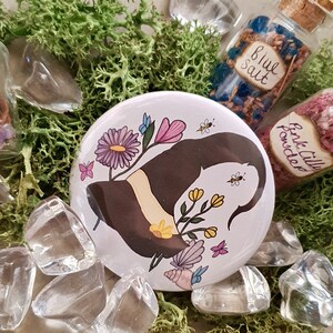 May include: A white round button featuring a black witch's hat with a white brim, surrounded by purple, pink, and yellow flowers. The button is surrounded by clear glass stones and moss.