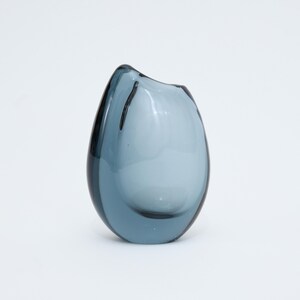 Könnte beinhalten: Eine tropfenförmige, blau-graue Glasvase. Die Vase hat eine glatte, glänzende Oberfläche und eine einzigartige, asymmetrische Öffnung. Das Glas ist durchscheinend und lässt Licht durch. Ein dekorativer Artikel.