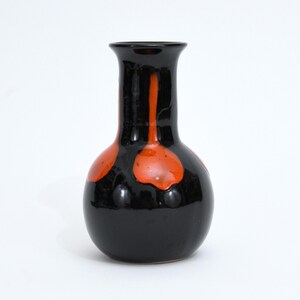 Könnte beinhalten: Eine schwarze Keramikvase mit rundem Körper und schmalem Hals. Die Vase weist leuchtend orangefarbene abstrakte Designs auf. Die glänzende Oberfläche verstärkt den Kontrast zwischen Schwarz und Orange und erzeugt einen auffälligen visuellen Reiz. Ein dekoratives Stück für die Inneneinrichtung.