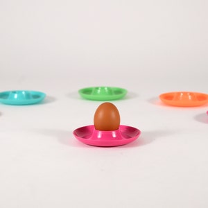 May include: A brown egg sits in a pink egg cup, surrounded by five empty egg cups in various colours: turquoise, green, orange, and pink.