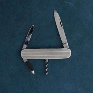 Può includere: Un coltello tascabile argentato con più lame e un cavatappi, su sfondo blu scuro. L'impugnatura del coltello ha un design strutturato e modellato. Una lama è aperta, mostrando il testo "Solingen Germany".