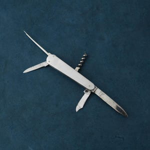 May include: A silver multi-tool with various open blades and tools, including a corkscrew, a small knife, and a pointed awl. The tool has a rectangular body against a dark blue background.