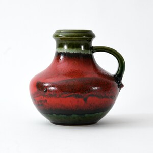 Op de afbeelding: Een keramische kan met een afgeronde body en een handvat. De kan heeft een kleurenschema van rood, groen en zwart. De bovenkant en het handvat zijn groen, terwijl de body overgaat van rood naar zwart en groen. De kan is waarschijnlijk voor decoratief gebruik.