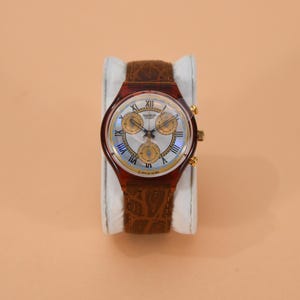 Può includere: Un orologio cronografo Swatch con cinturino in pelle marrone. Il quadrante è bianco con sotto-quadranti color oro e numeri romani. La cassa è in plastica trasparente rosso-marrone.