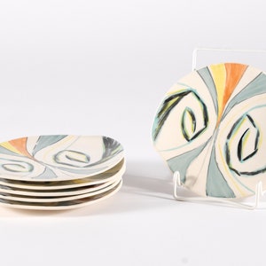 Può includere: Set di sei piatti in ceramica con un design astratto a spirale in tonalità di blu, verde, giallo e arancio. I piatti sono impilati uno sopra l'altro.