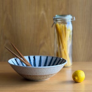 May include: A speckled ceramic bowl with dark blue vertical stripes, containing two wooden chopsticks. Beside the bowl sits a clear glass jar filled with long pasta strands and a single bright yellow lemon on the surface.