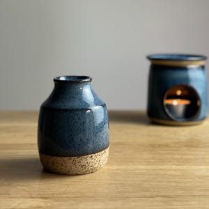 May include: A small, blue ceramic vase with a speckled finish and a tan base. A blue ceramic oil warmer with a round opening and a lit candle inside is in the background. Both items are on a wooden surface.