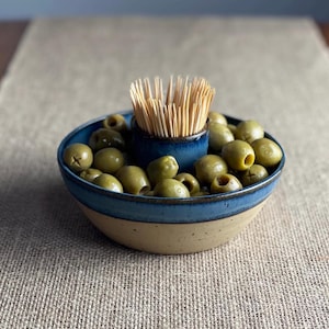 May include: A ceramic olive and toothpick dish with a blue and beige glaze. The dish has a central compartment for toothpicks and a larger surrounding area for olives.