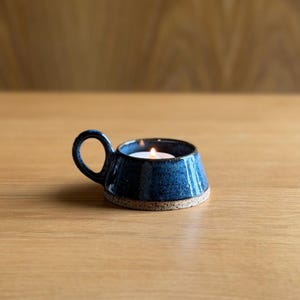 May include: A small, round, dark blue ceramic candle holder with a speckled finish and a single side handle. The base is unglazed, showing a light tan, textured clay. A lit white tea light candle sits inside the centre of the holder.