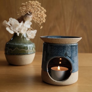 May include: A ceramic wax warmer with a blue glazed top and a speckled beige base, featuring a circular opening containing a lit tea light candle. Beside it sits a matching ceramic vase filled with dried white flowers and seed pods.