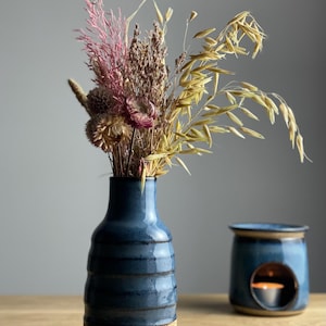 May include: A blue ceramic vase with a beige base holds a bouquet of dried flowers in shades of pink, brown, and yellow. A matching blue oil warmer with a lit candle sits nearby. The vase is approximately 15 cm tall.
