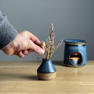 May include: A small, blue ceramic vase with a speckled tan base holds dried flowers. A hand is placing a dried seed pod into the vase. A blue oil warmer with a lit candle sits nearby on a wooden surface.