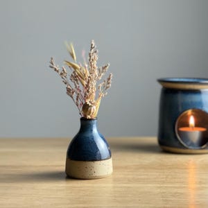 May include: A small ceramic vase with a dark blue top and a beige base, containing dried flowers. The vase sits on a wooden surface. A blue oil burner with a lit candle is in the background.