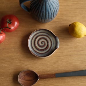 May include: A round ceramic spoon rest featuring a brown and beige spiral pattern, positioned near a blue ribbed pitcher, two red tomatoes, a yellow lemon, and a wooden spoon with a dark grey handle on a light brown wooden surface.