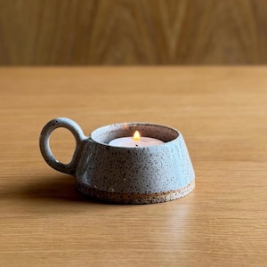 May include: A speckled light grey ceramic candle holder with a single circular handle. The bowl-shaped vessel features a tapered design and a rough, textured base. A small white tea light candle is placed inside, with a lit flame visible.